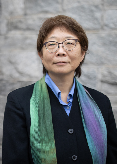 A Japanese woman stands in front of a stone wall in a colorful scarf and glasses