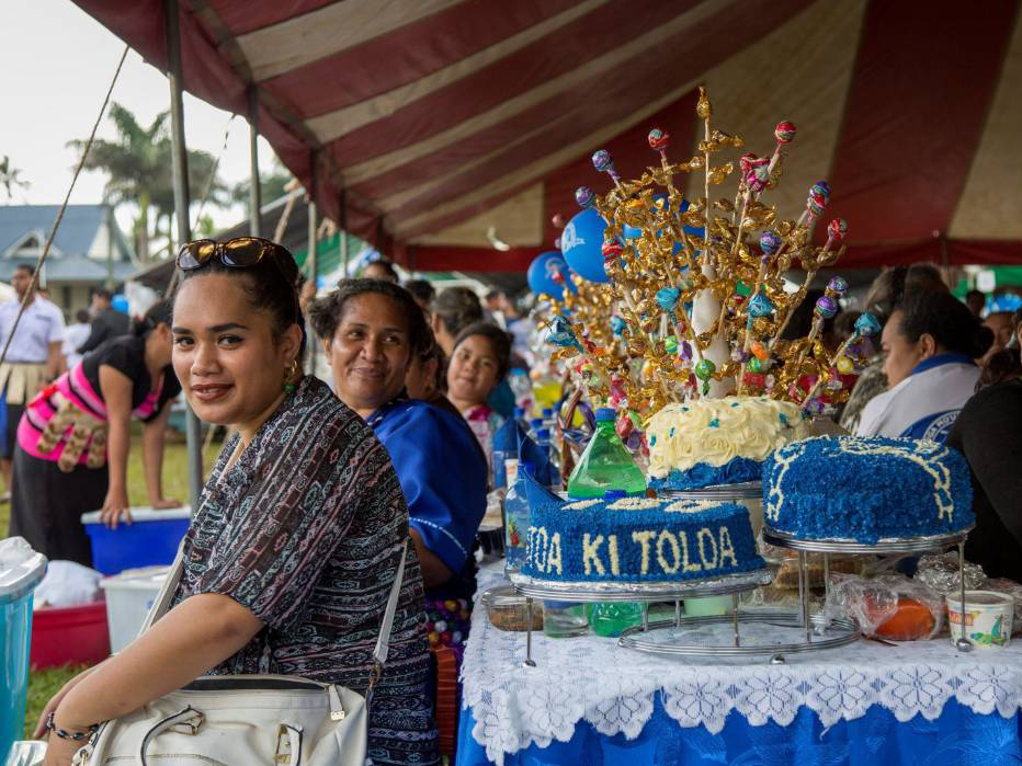 Film still from For My Father's Kingdom. Women in Tonga sit at a table alongside a festive cake. 