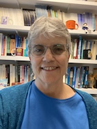 Man in round glasses, wearing a suit and tie, standing in front of a sun-dappled campus