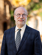 Man in round glasses, wearing a suit and tie, standing in front of a sun-dappled campus