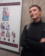 Man in round glasses, wearing a suit and tie, standing in front of a sun-dappled campus