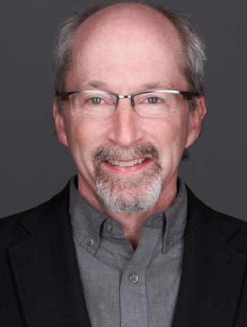 Man in square glasses with a beard, wearing a grey button-down shirt and a black jacket, smiling in front of a grey background