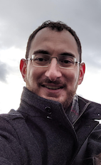 Man in round glasses, wearing a suit and tie, standing in front of a sun-dappled campus