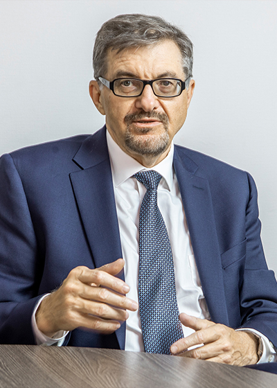 Seated man in suit, tie, and glasses, gesturing with his hands