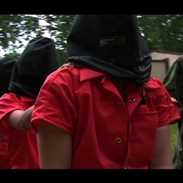 Film still from Operation Atropos. Women in orange jump suits with black hoods over their heads. 