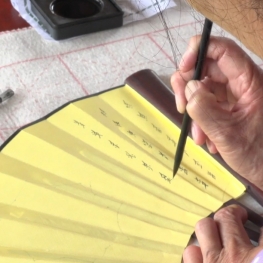Film still from Her Words, woman writing calligraphy on a yellow hand fan